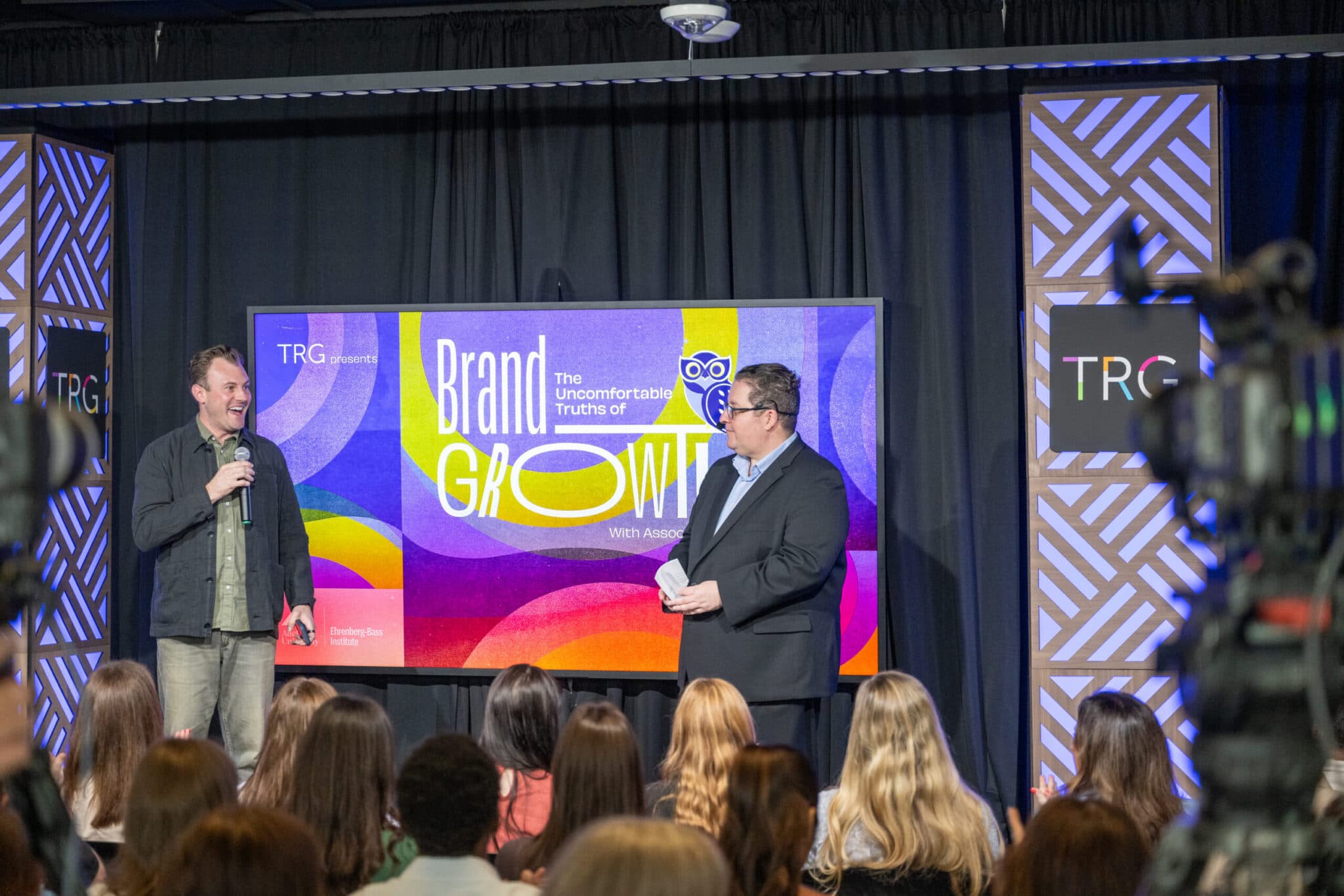 A speaker presenting on stage with a microphone beside a host, in front of the colorful “Brand Growth” screen