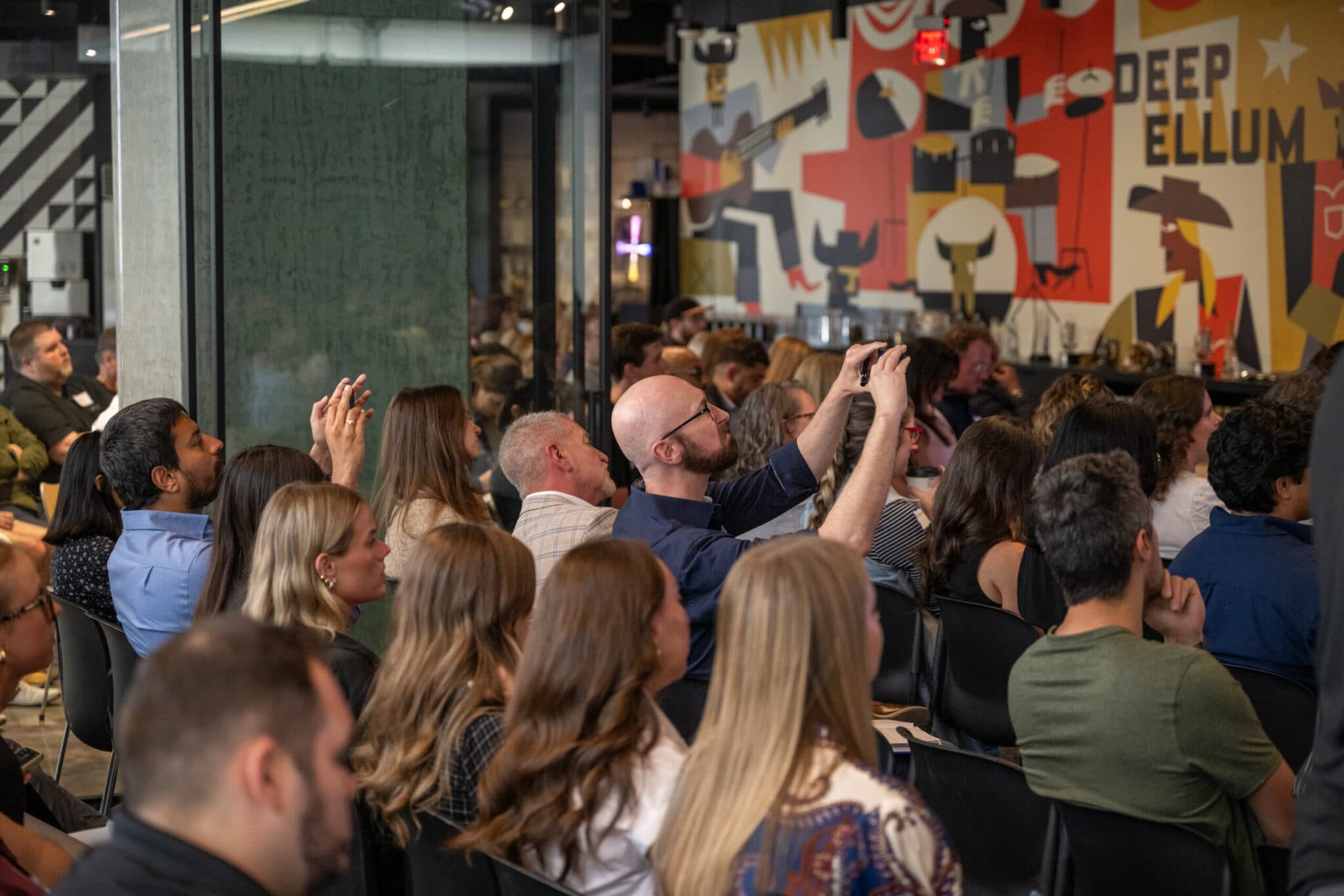 Attendees seated in the audience, one raising a phone to photograph the stage, beside a bold “Deep Ellum” mural