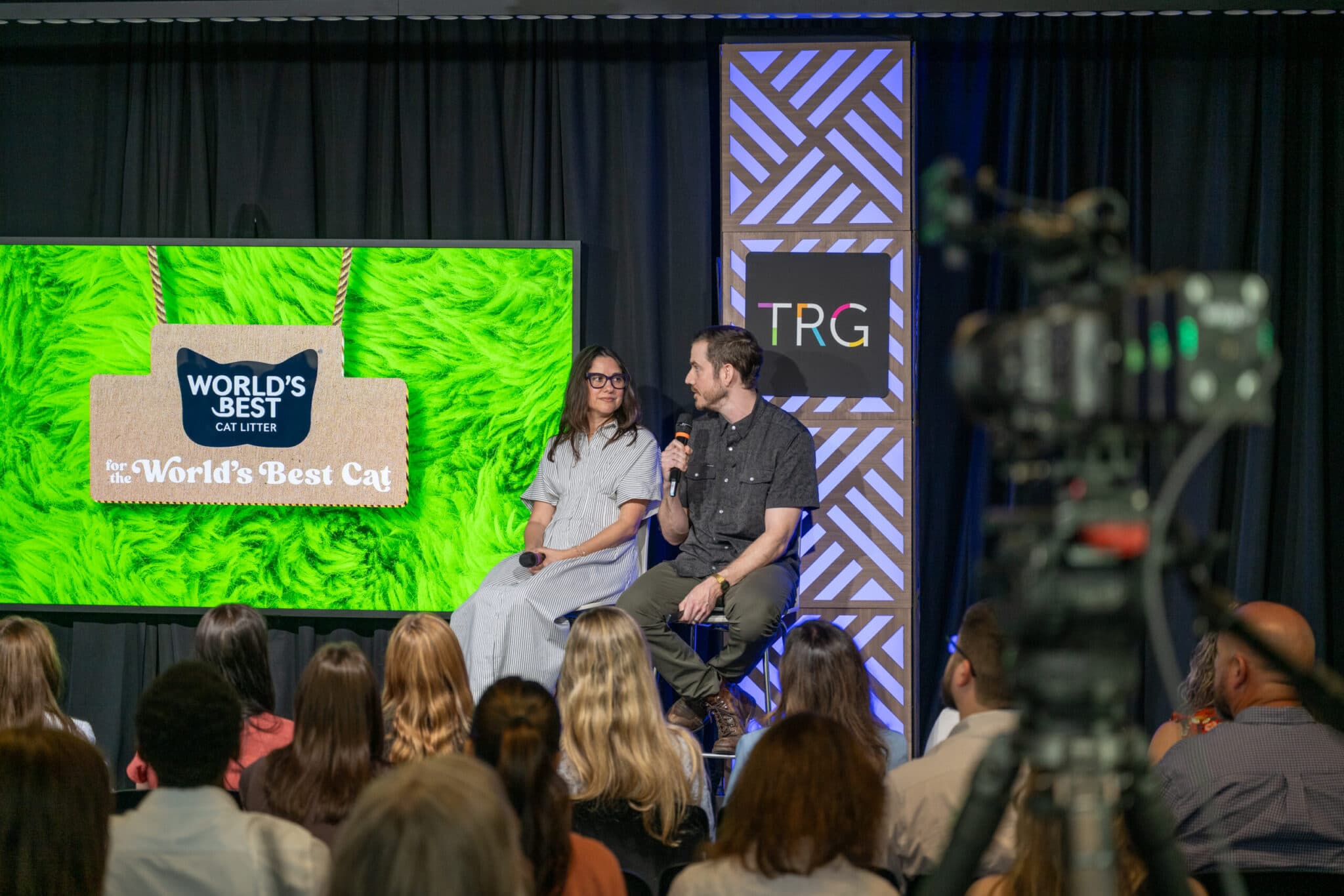 Two speakers in conversation on stage with a “World’s Best Cat Litter” presentation on the screen behind them