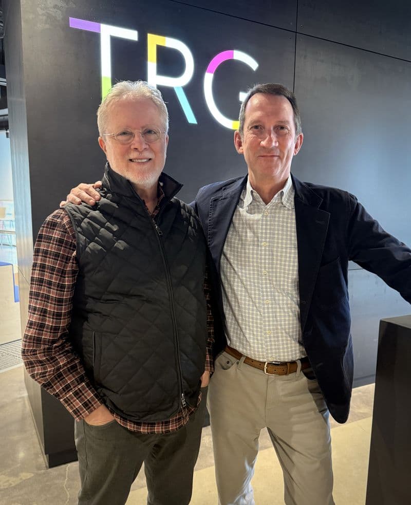 Glenn Dady and Pete Lempert standing together in front of the illuminated TRG logo wall